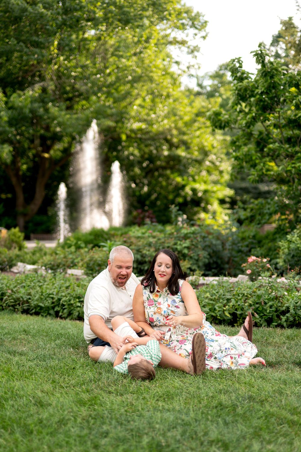 Couple sitting on green grass in a garden park with a fountain and lush trees in the background on a sunny day.