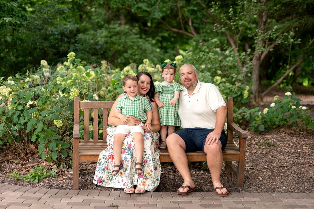 Family of four smiling on a wooden bench in a lush garden, wearing coordinating green and white outfits.