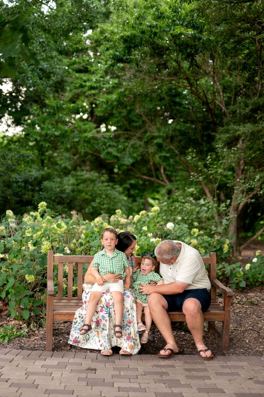 Family of four sitting together on a wooden bench surrounded by blooming flowers in a garden park.