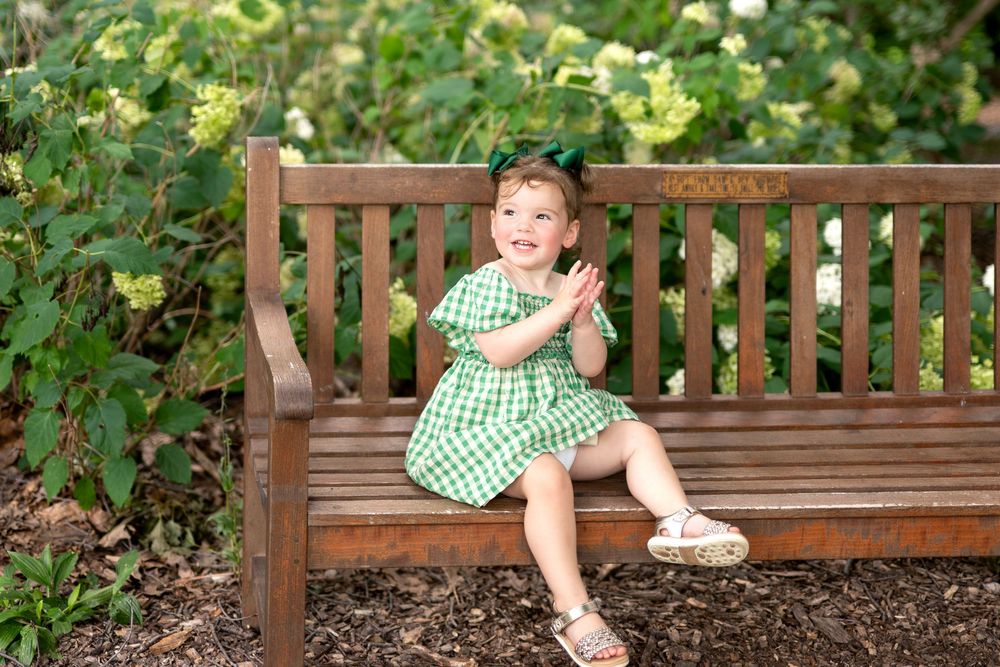 Adorable toddler girl in green gingham dress sitting on wooden bench clapping in garden.