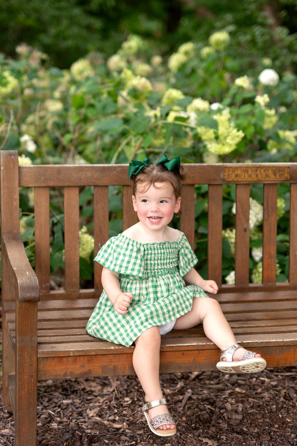 Smiling toddler girl in green gingham dress and bow sits on wooden bench surrounded by lush garden greenery.