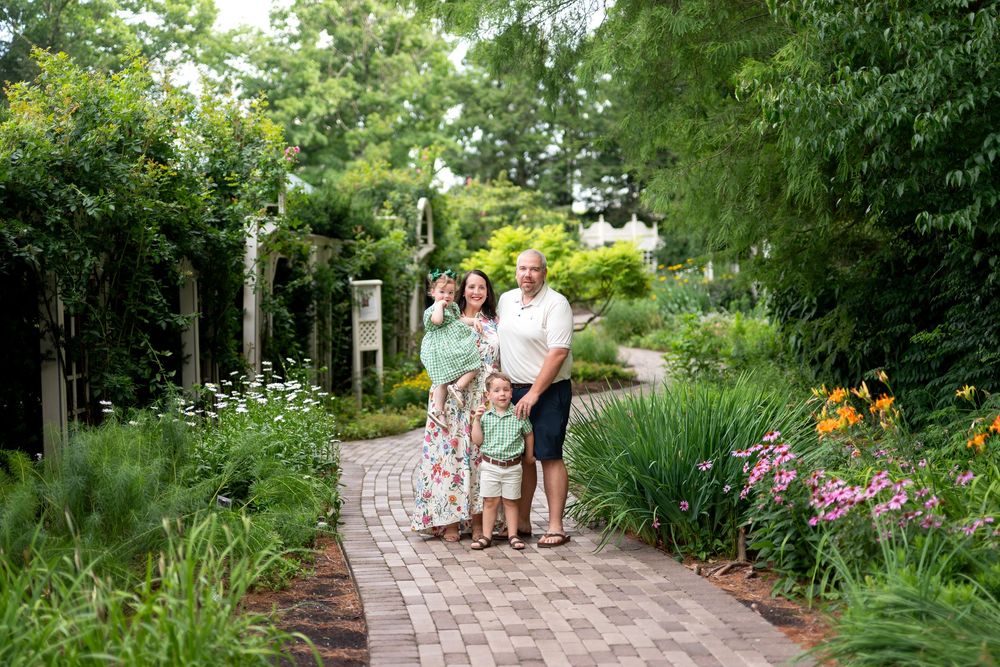 Family of four posing on a brick garden path surrounded by lush greenery and colorful flowers on a sunny day.