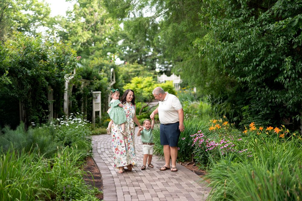Family of four walking on a brick garden path surrounded by lush greenery and colorful flowers on a sunny day.