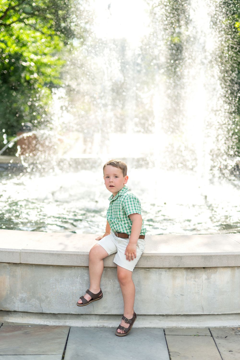 Young boy in green plaid shirt and white shorts sitting on fountain edge in sunny park.