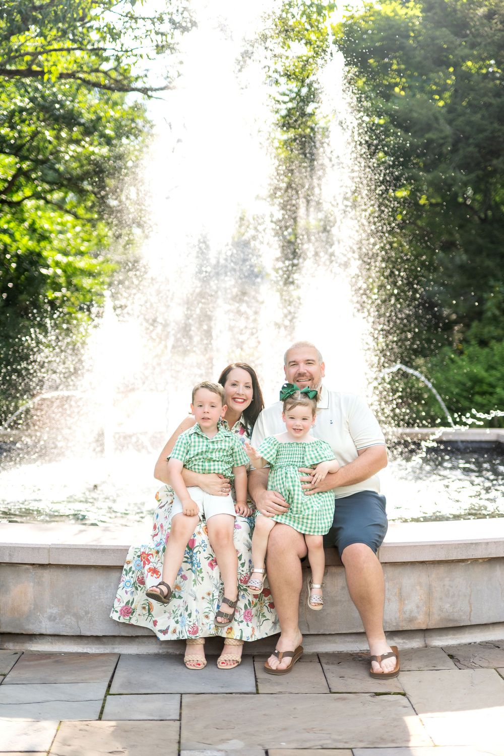 Family of four in matching green outfits sitting by a fountain outdoors, smiling together on a sunny day.