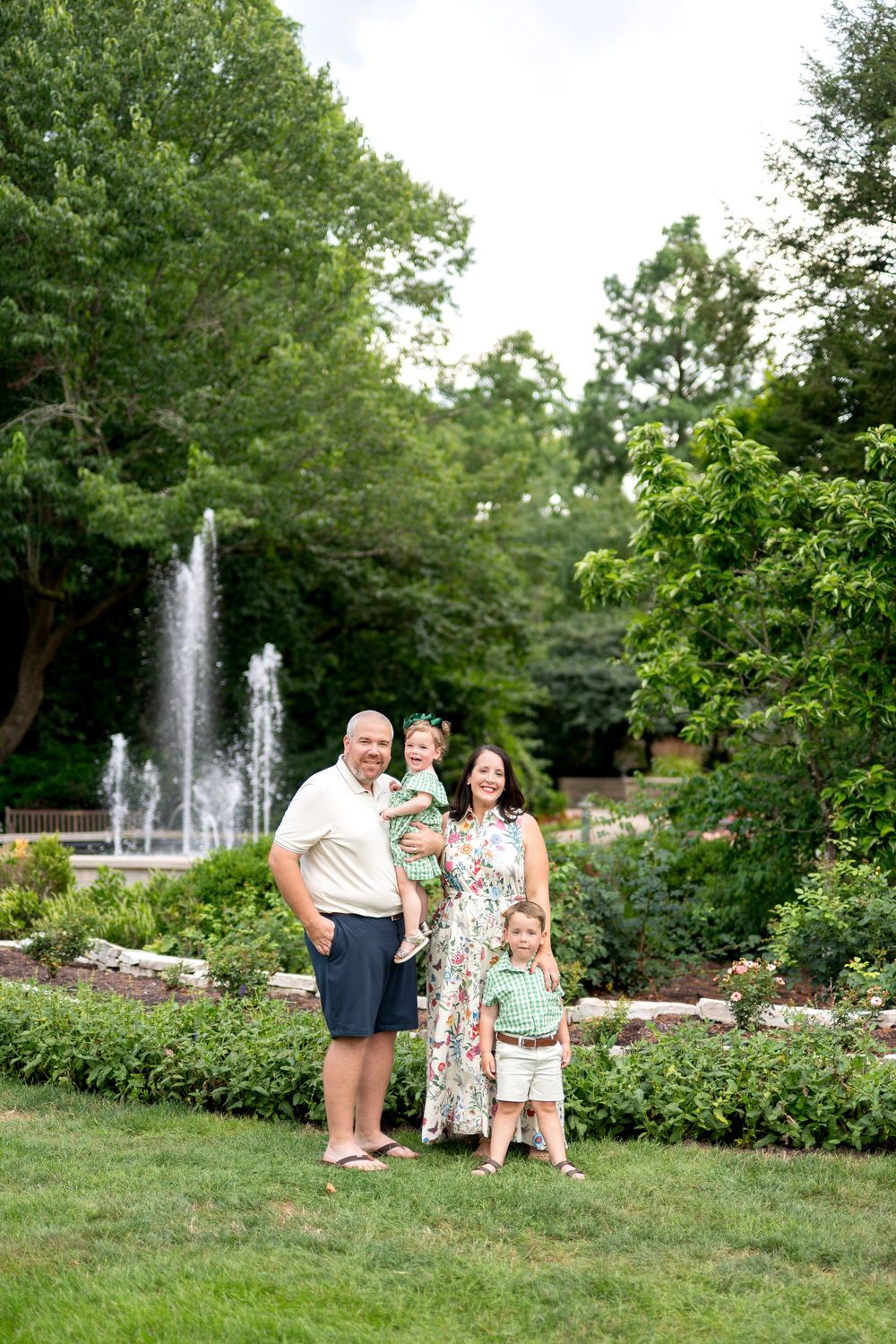 Family of four posing in a lush garden park with a fountain backdrop on a sunny summer day.