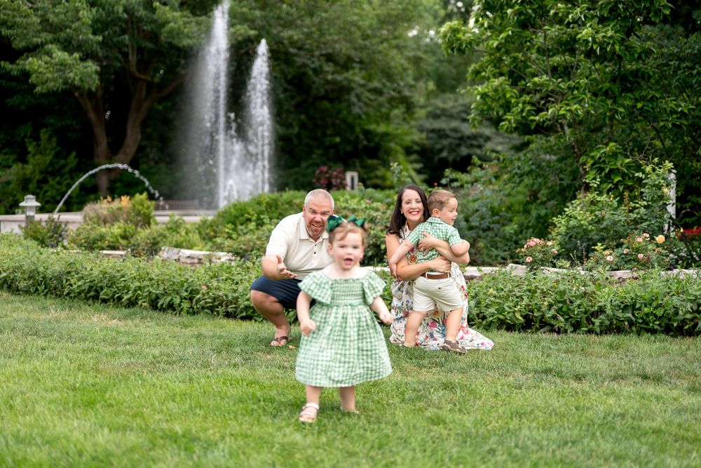 Family of four poses near a fountain in a lush green garden, with a toddler girl in a green dress running toward the camera.