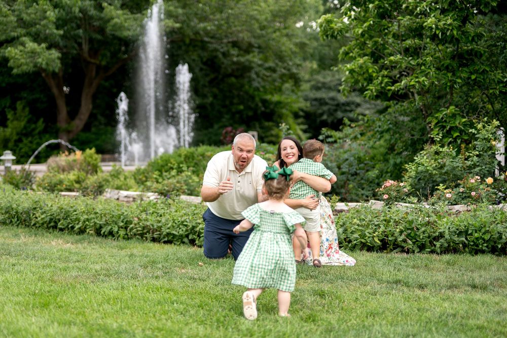 Family playing in a lush green park with a fountain, parents embracing children in matching green outfits outdoors.