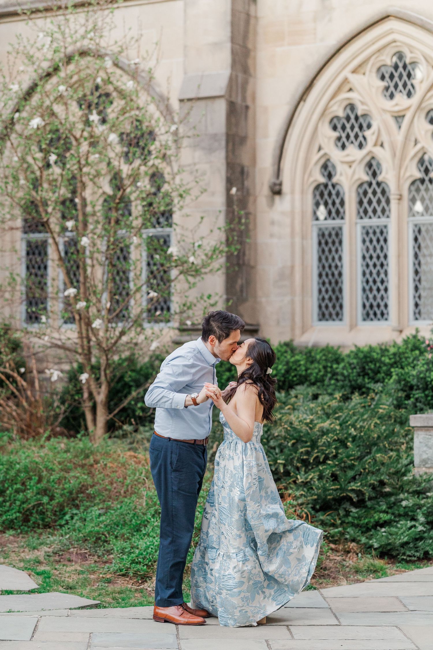 Couple sharing a romantic kiss in a light blue floral gown outside a Gothic stone chapel with arched windows.