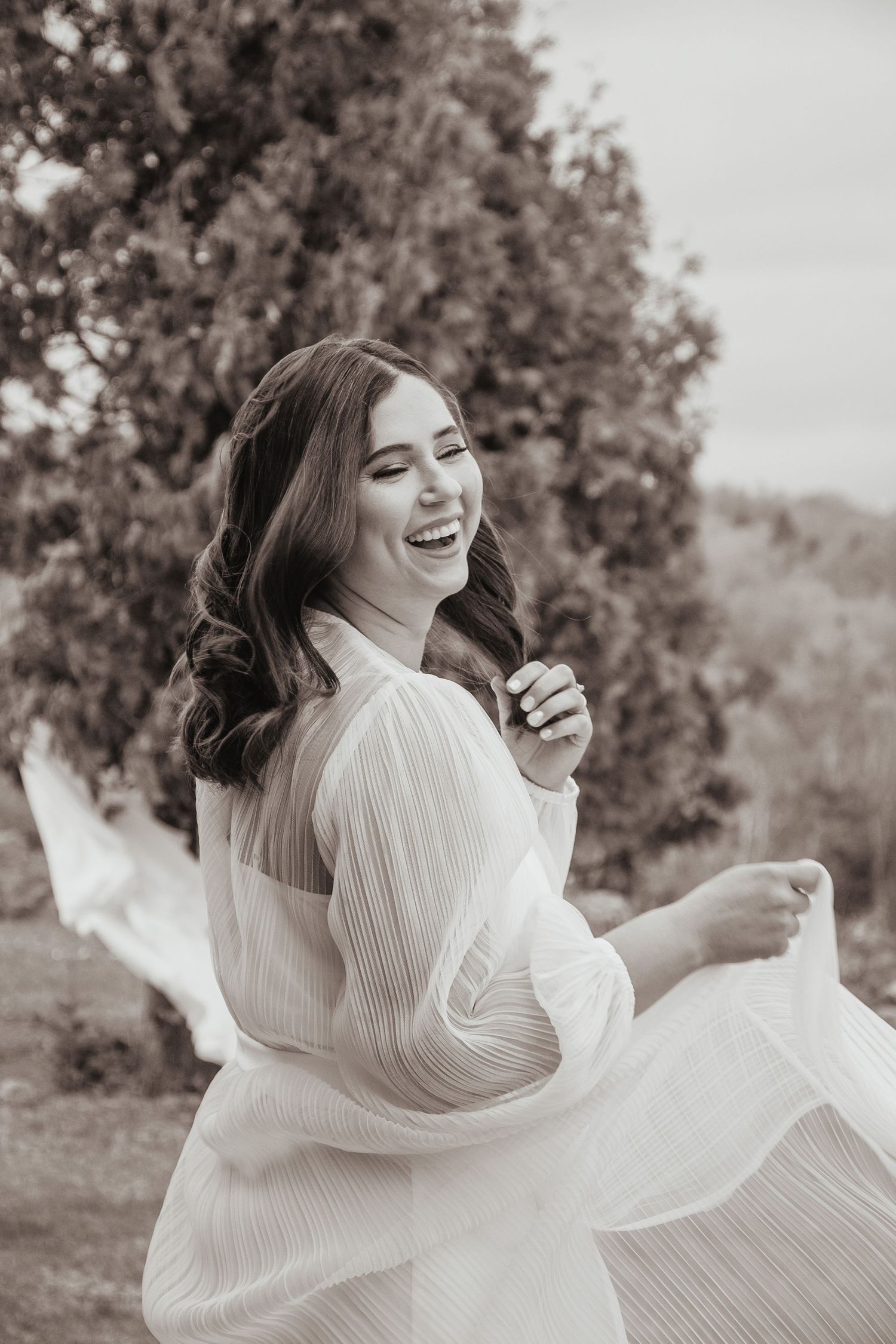 A joyful bride in a flowing white gown laughs outdoors in a black and white portrait with trees in the background.
