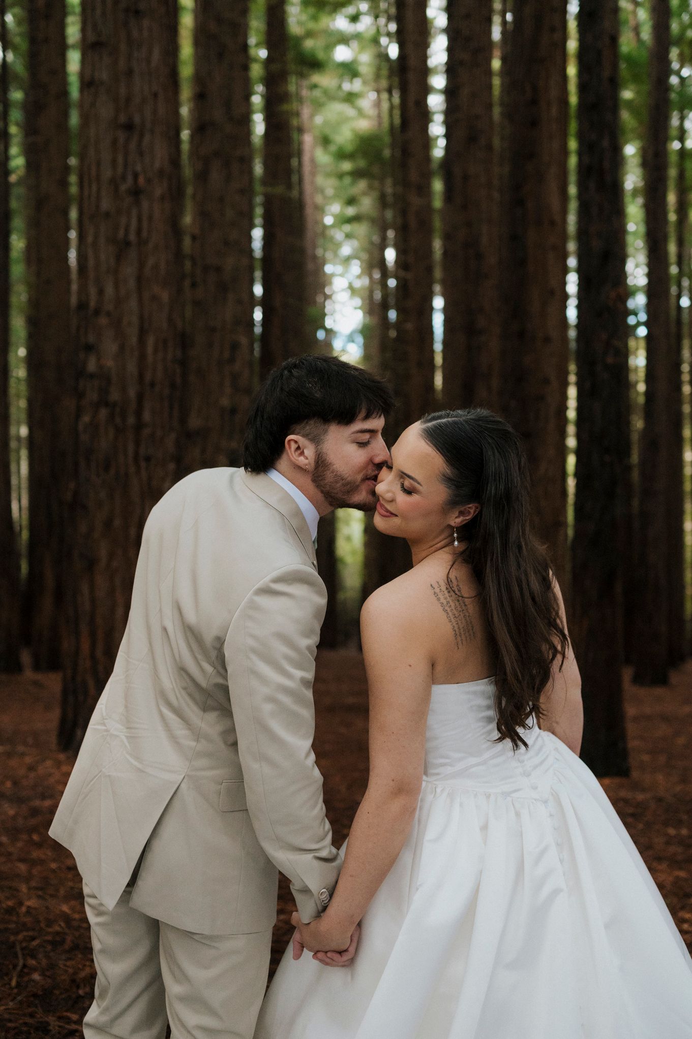 A bride and groom share a tender kiss while holding hands in a tall redwood forest during their wedding day.