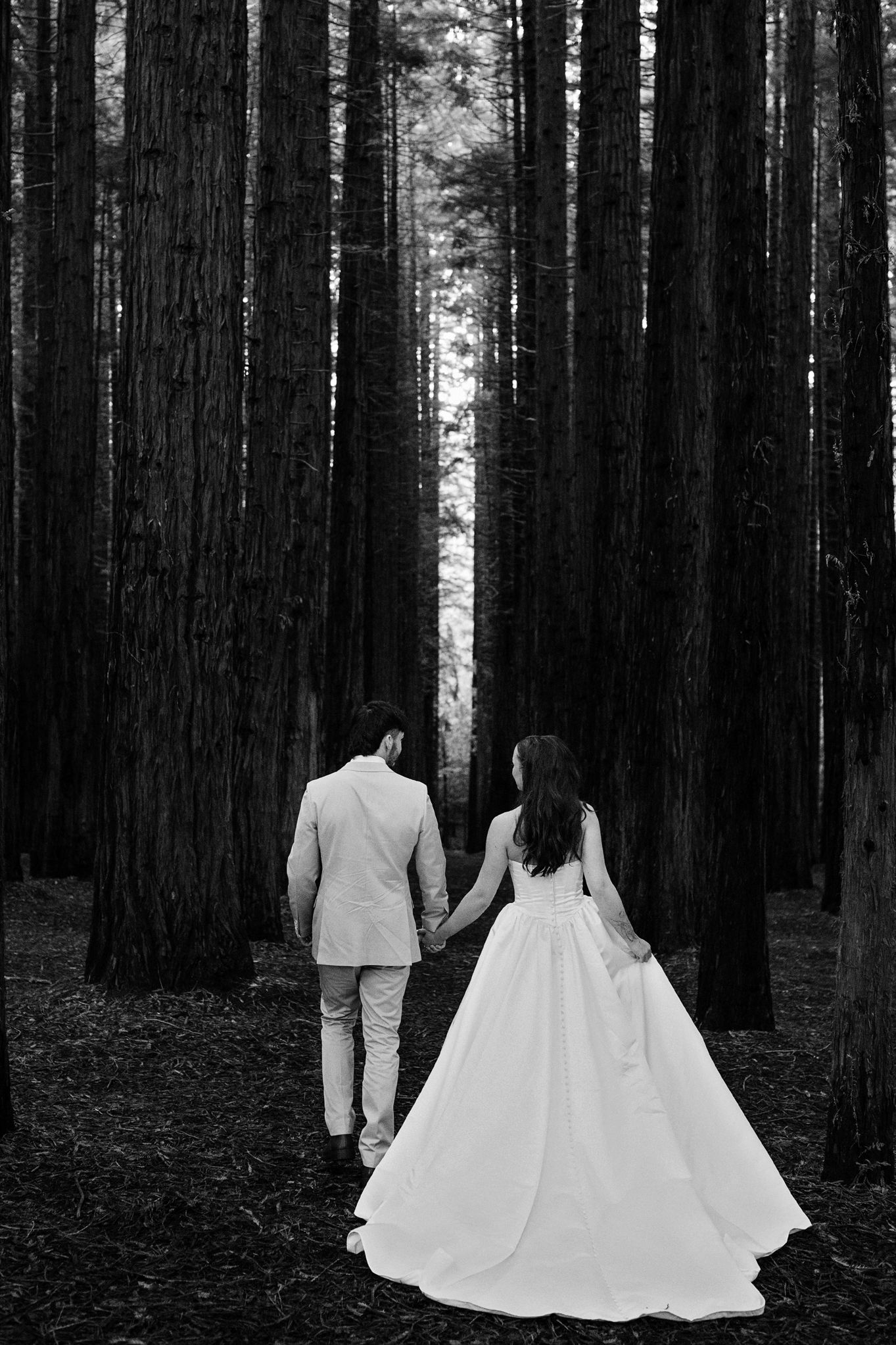 Black and white photo of bride and groom holding hands walking through tall forest trees in wedding attire.