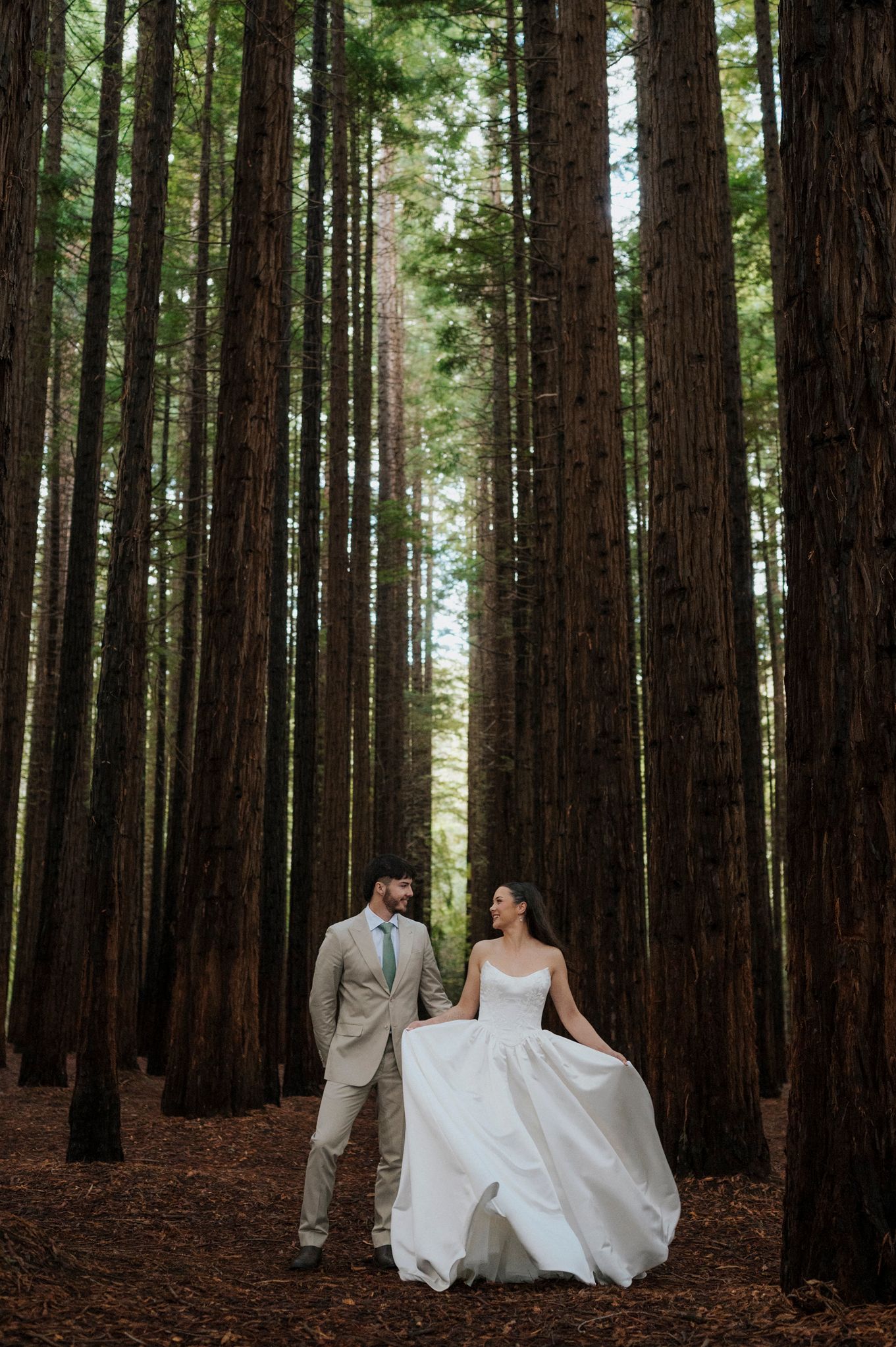 Bride and groom walking hand in hand through tall redwood forest, bride in flowing white gown, groom in tan suit.