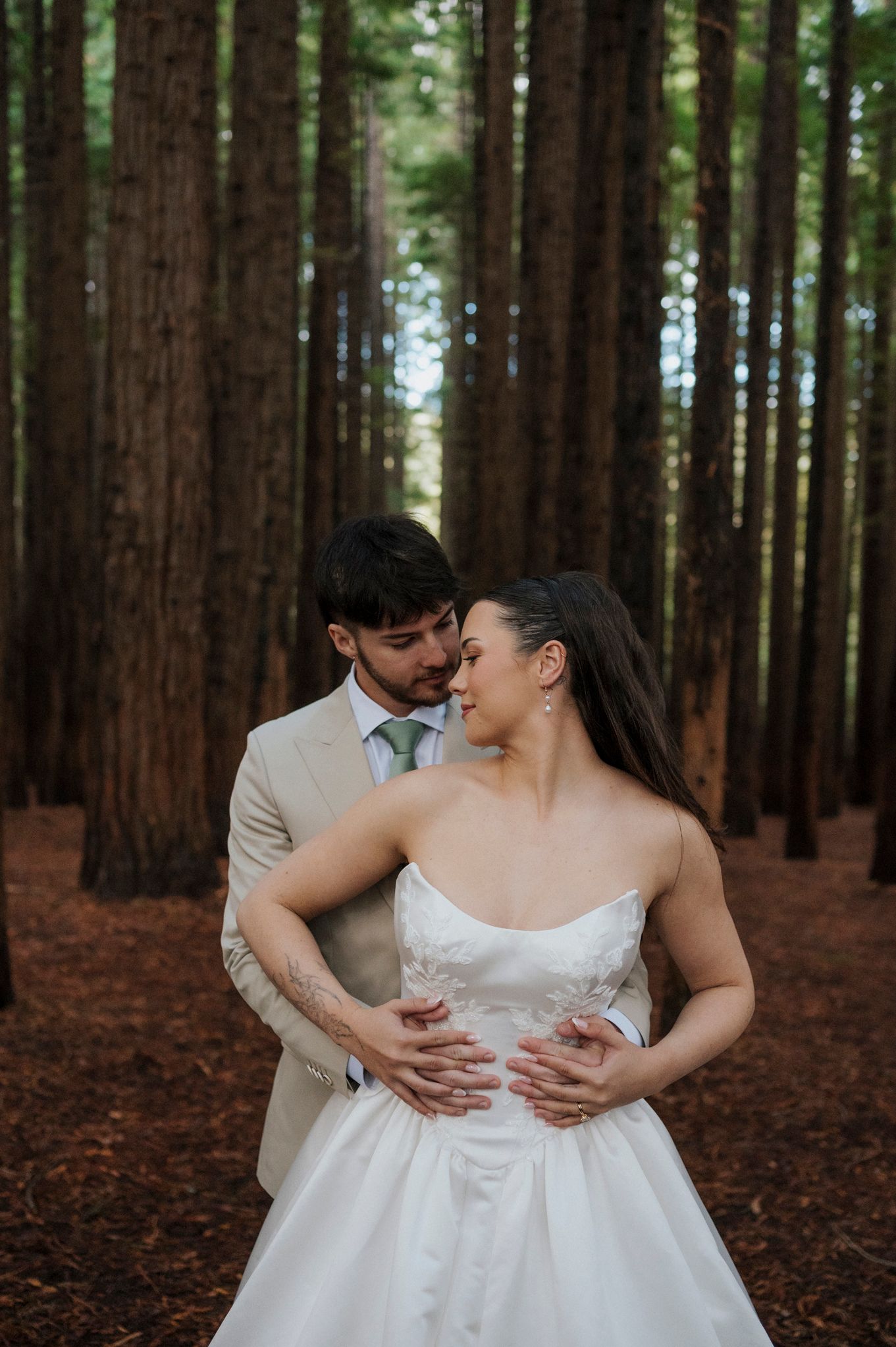 Bride and groom embrace romantically in a tall redwood forest, she in a strapless gown, he in a beige suit with green tie.