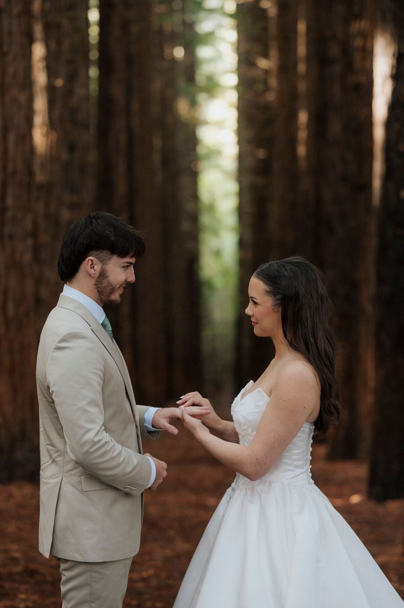 Bride placing wedding ring on groom's finger in a romantic forest setting, both dressed in elegant wedding attire.
