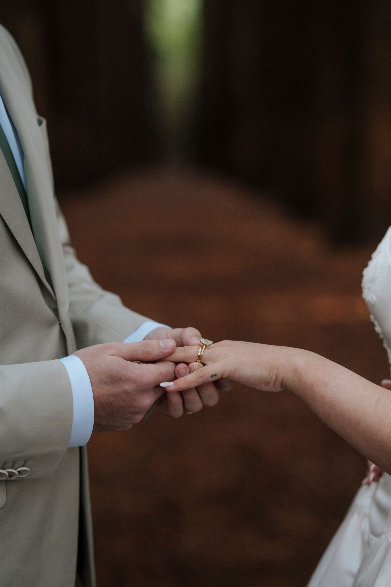 Groom placing a gold wedding ring on bride's finger during outdoor ceremony, with blurred forest path background.