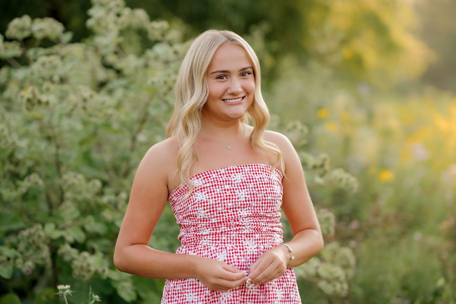 Young blonde woman smiling in pink floral strapless dress outdoors among green and yellow wildflowers.