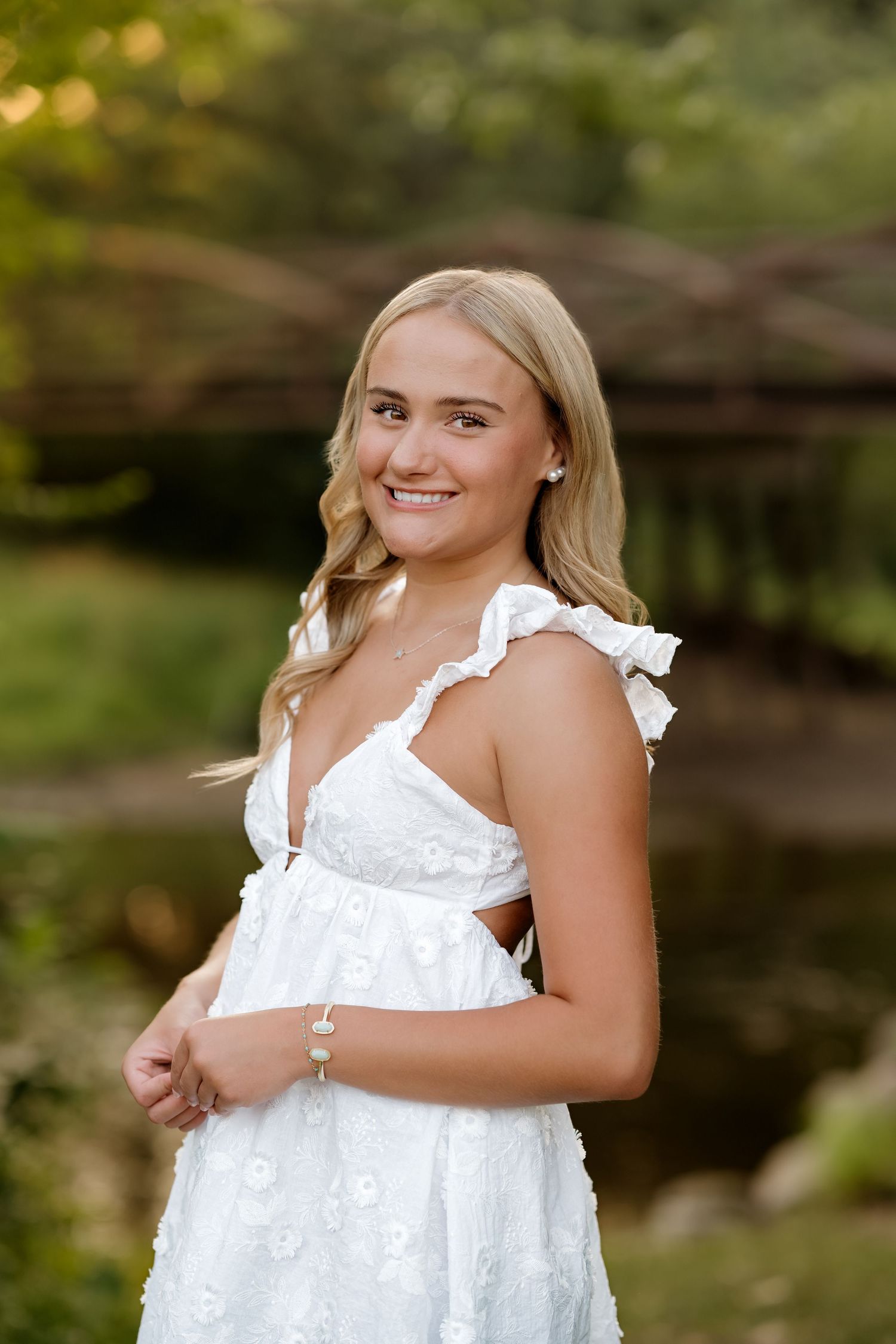 Young blonde woman smiling in white floral dress outdoors near a bridge surrounded by lush greenery.