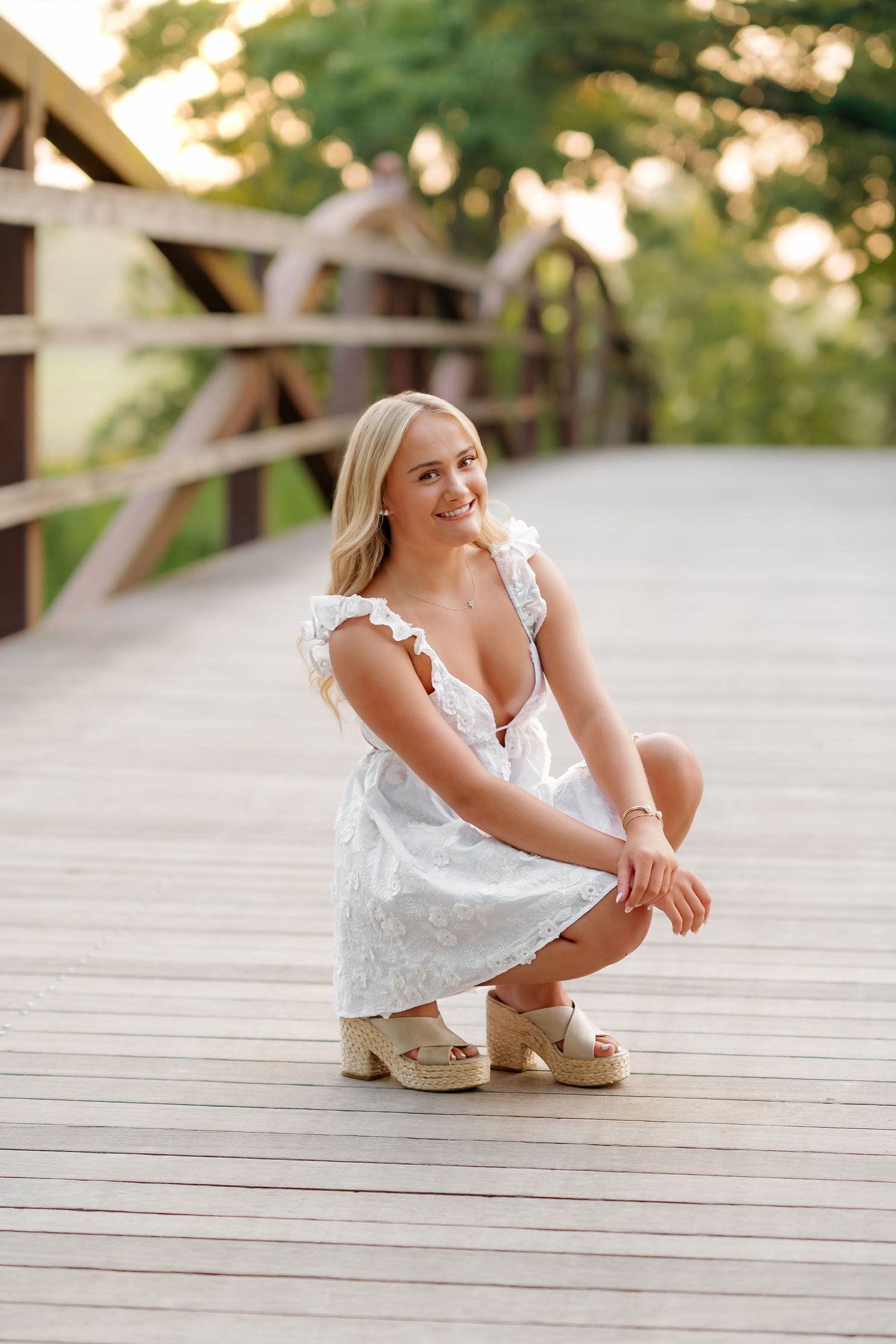 Young blonde woman in white dress crouching on wooden bridge, smiling outdoors in golden hour light.