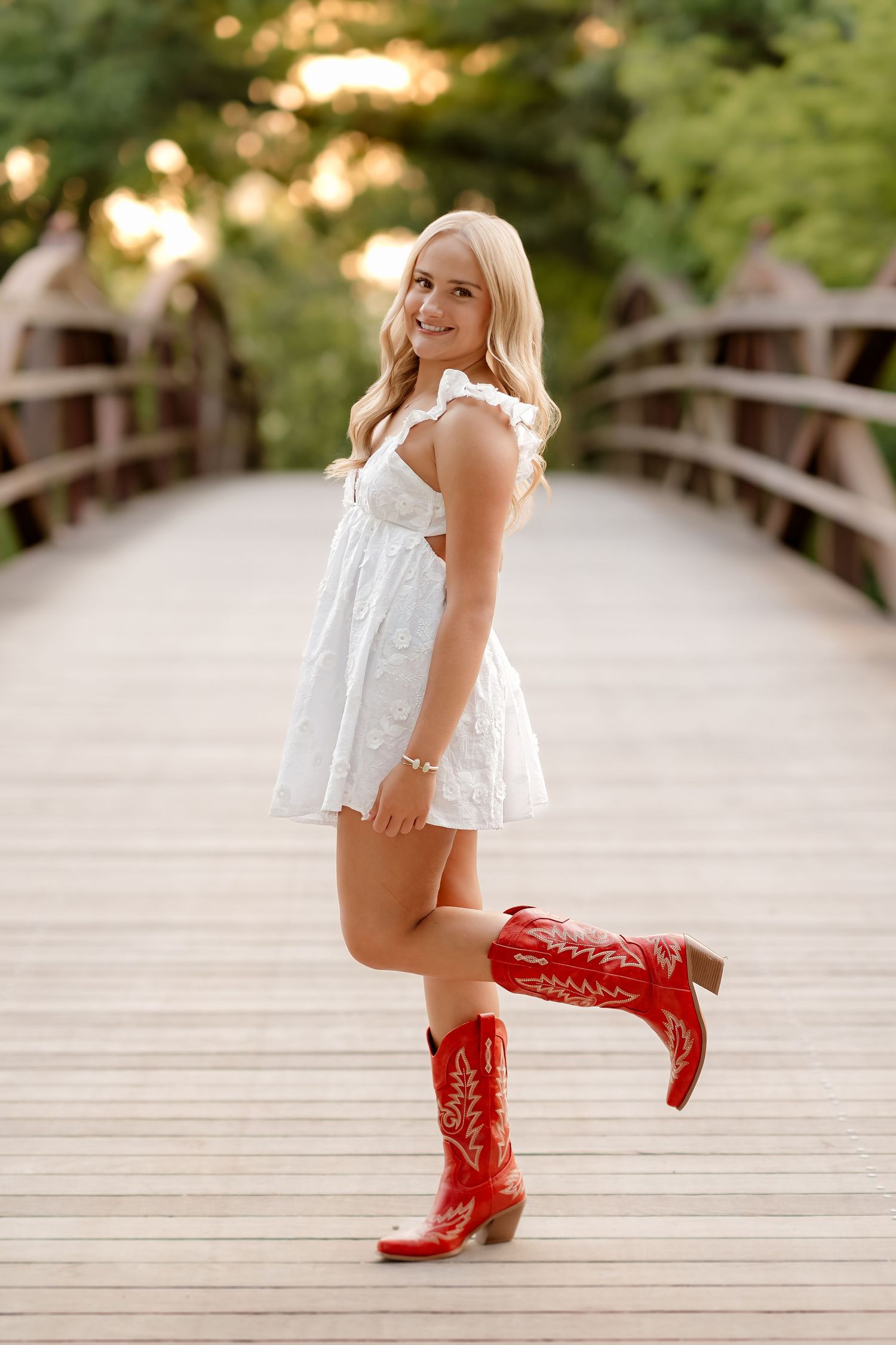 Young blonde woman in white dress and red cowboy boots posing on a wooden bridge outdoors.