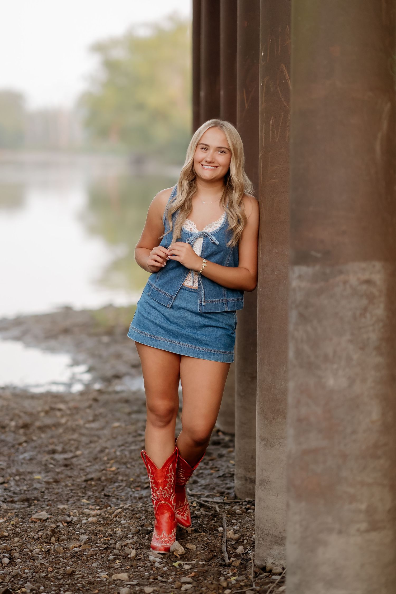 Young woman in denim mini dress and red cowboy boots leaning against a pillar near a river.