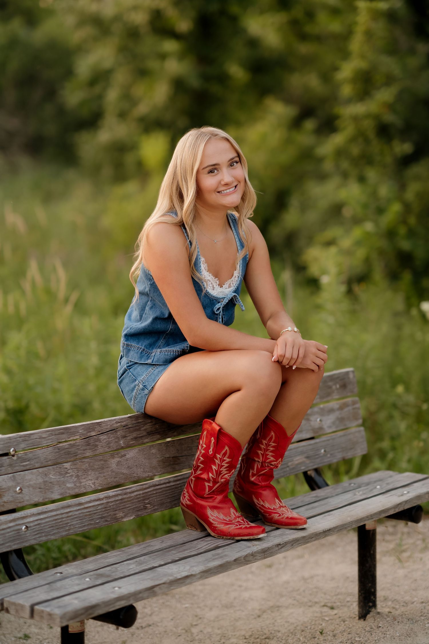Young blonde woman in denim romper and red cowboy boots sitting on a wooden bench outdoors.