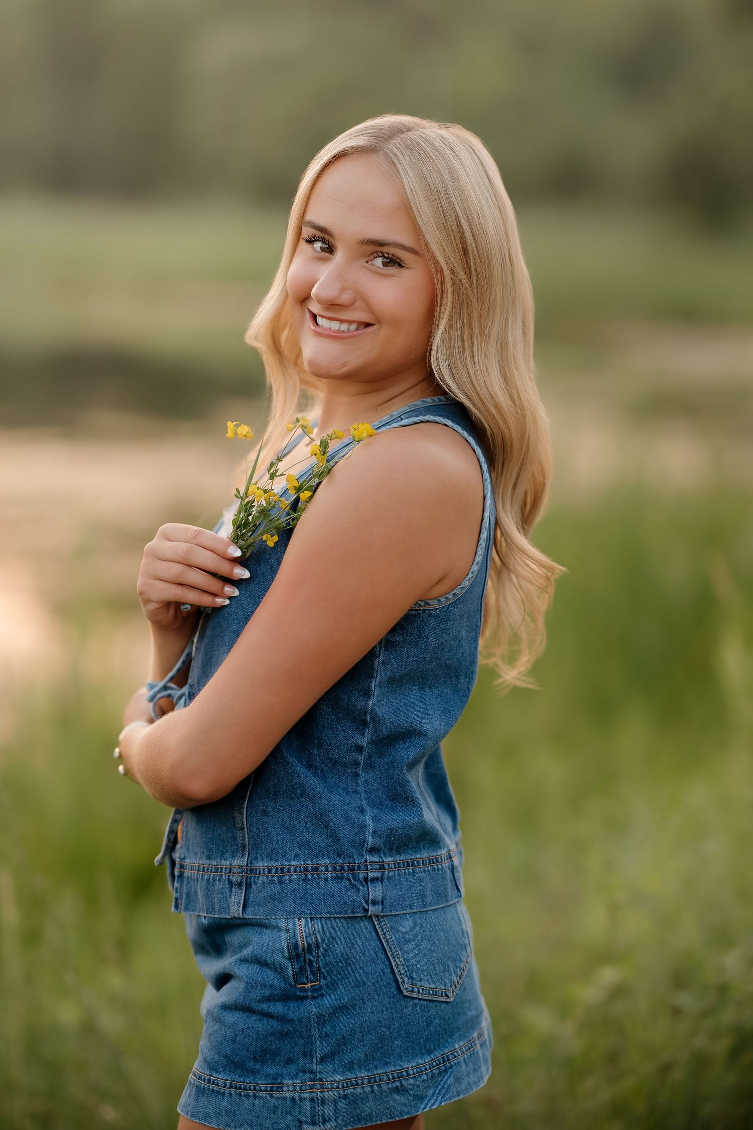 Smiling blonde teen girl in denim overalls holding wildflowers outdoors in a green field during golden hour.
