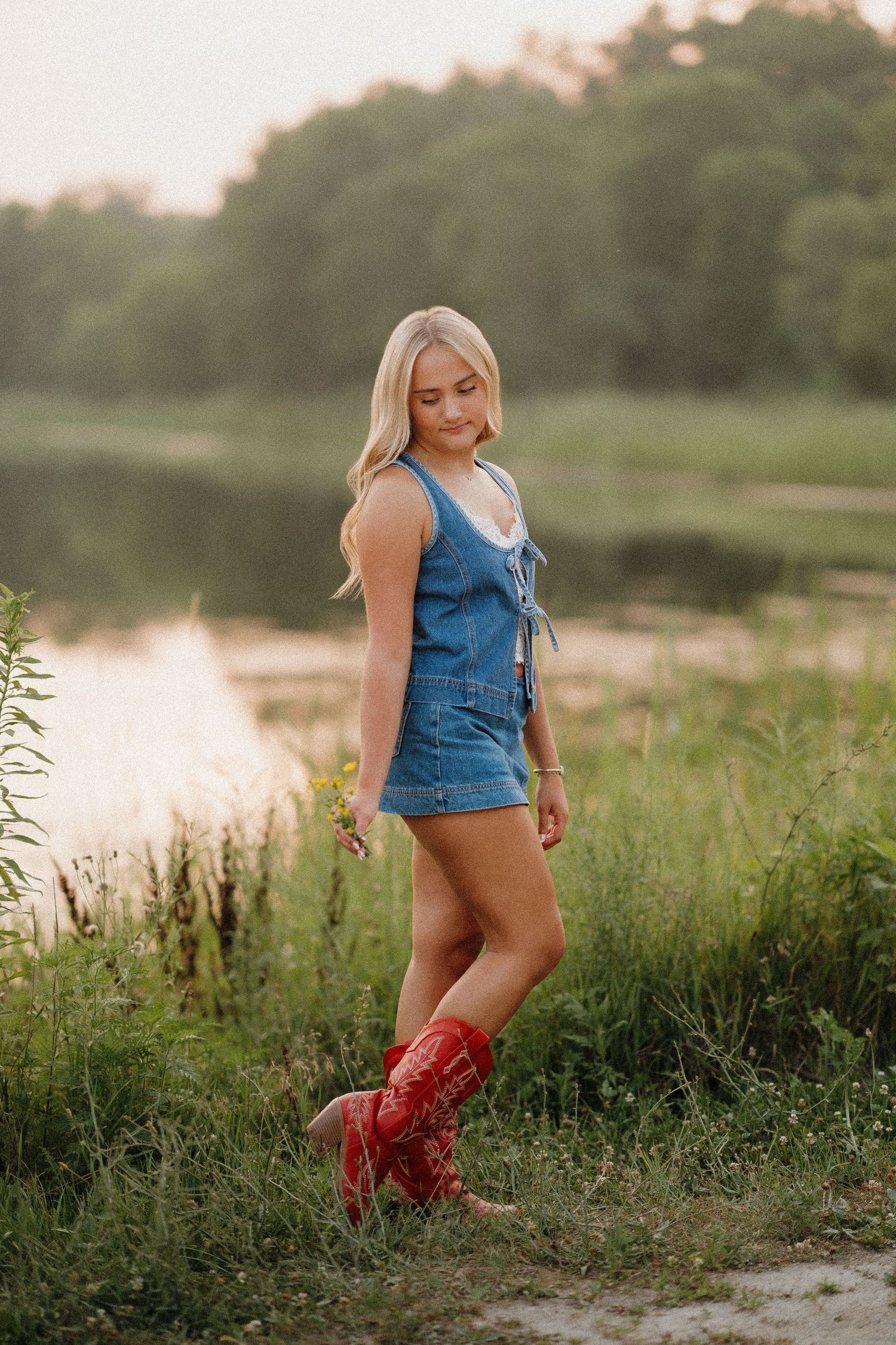 Blonde woman in denim romper and red cowboy boots standing by a lake in golden hour light.