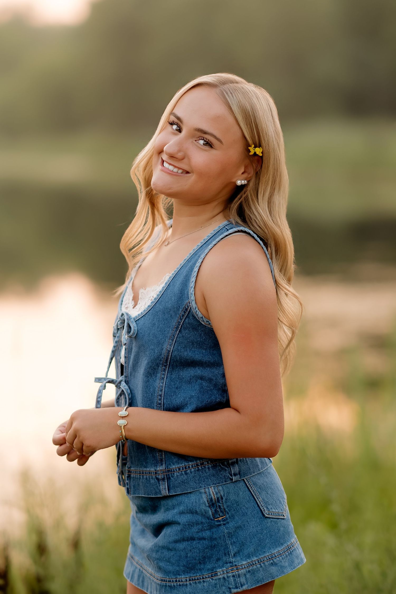 Smiling blonde teen girl in denim romper poses outdoors near a pond at golden hour with a yellow hair clip.