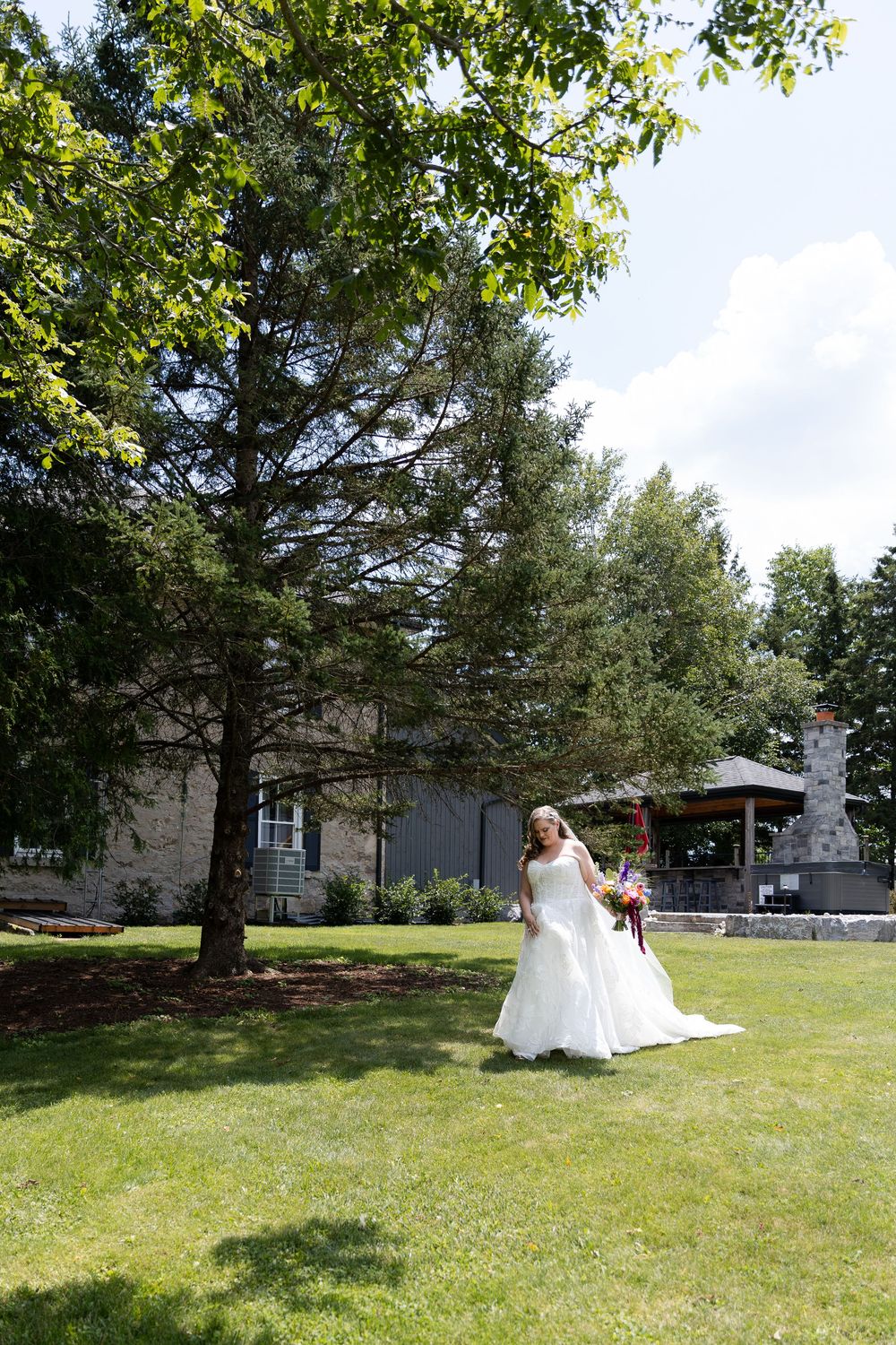 Bride in white gown holding bouquet stands on green lawn beside tall tree with stone building in background in Guelph, Ontario.