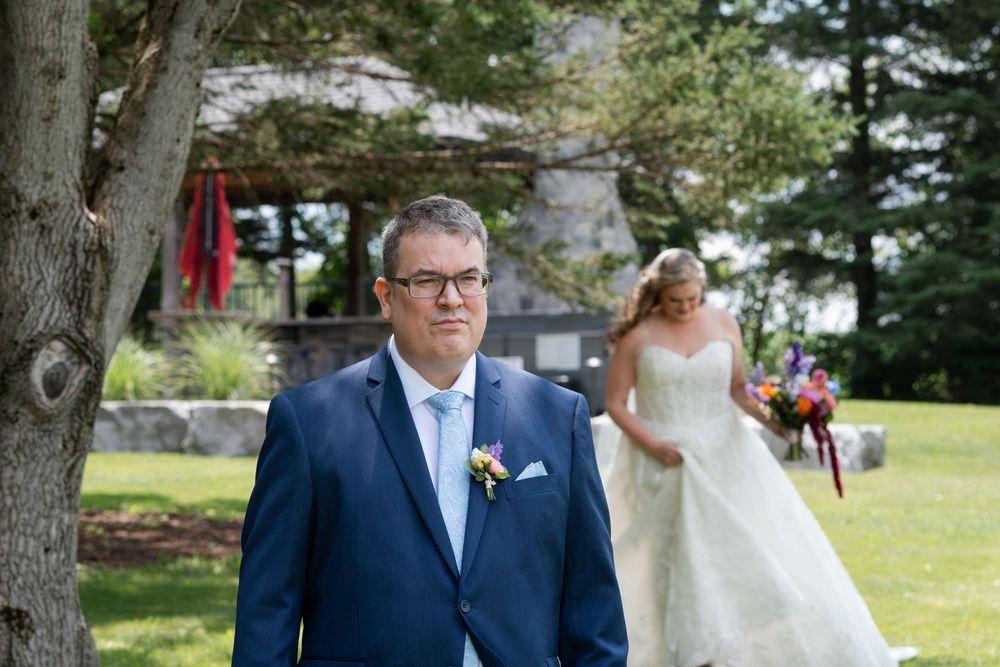 Groom in navy suit stands outdoors as bride in white gown approaches during a wedding first look ceremony in Guelph, Ontario.