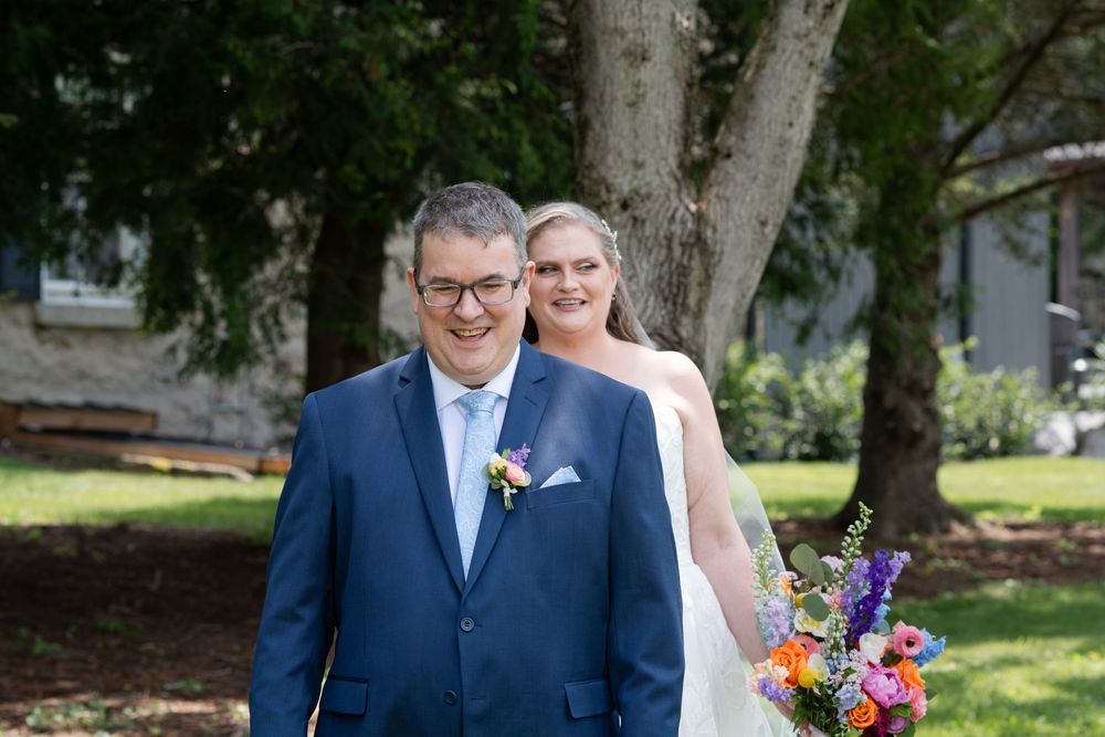 Bride holding colorful bouquet surprises groom in blue suit during outdoor wedding first look ceremony in Guelph, Ontario.