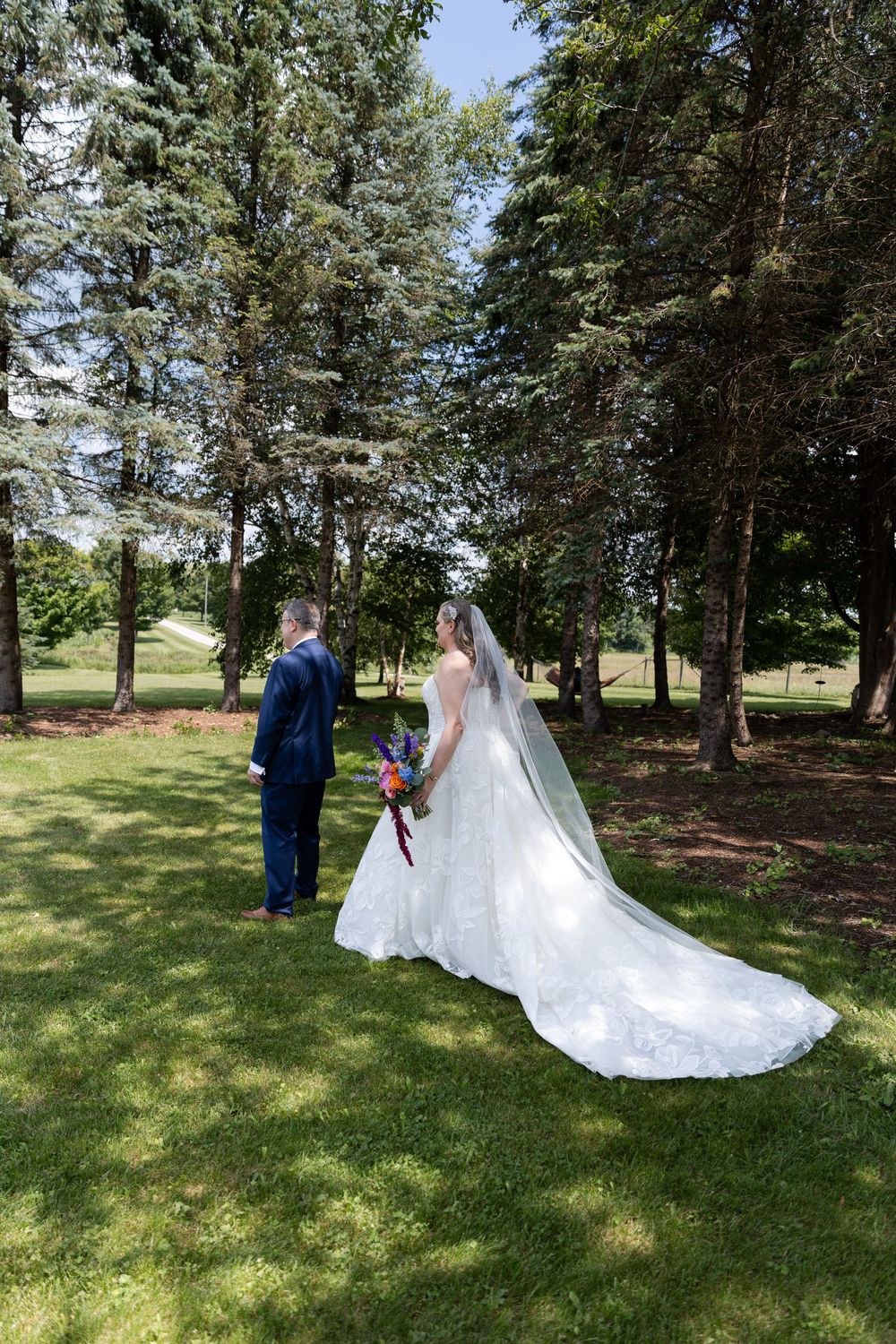 Bride in white gown with long veil holds bouquet beside groom in blue suit, standing on green grass among tall trees in Guelph, Ontario.