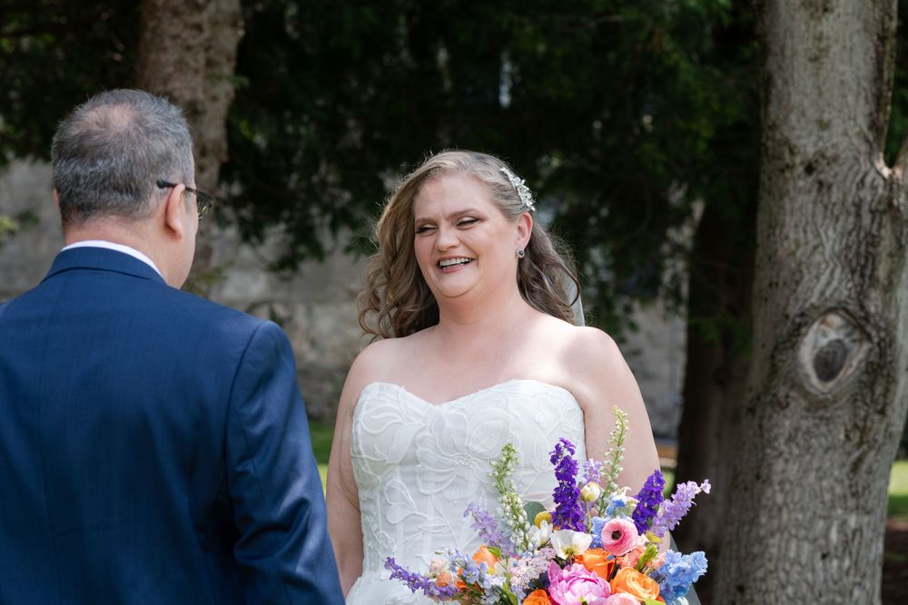 Smiling bride in strapless white gown holding colorful bouquet during outdoor wedding first look in Guelph, Ontario.