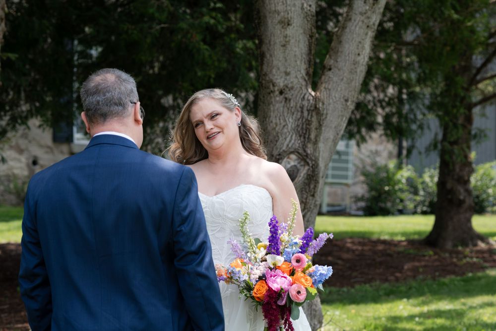 Bride in strapless gown holds colorful bouquet smiling at groom in navy suit outdoors near large tree in Guelph, Ontario.