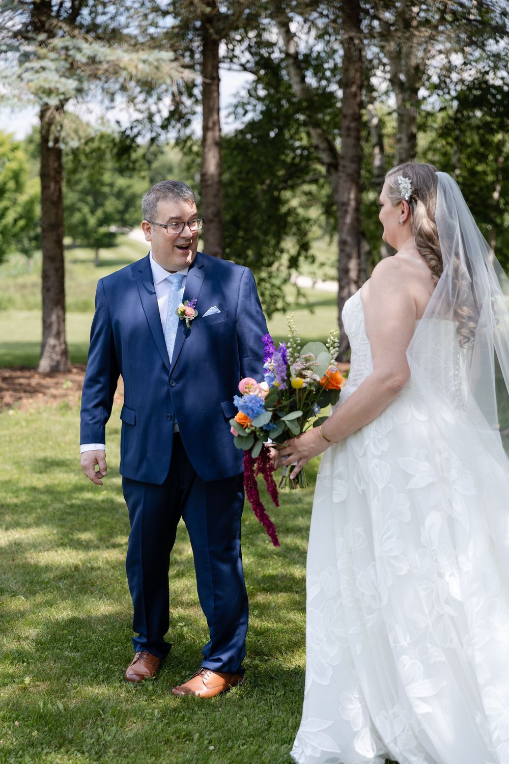 Groom in navy suit reacts with joy seeing bride in white gown holding colorful bouquet during first look outdoors in Guelph, Ontario.