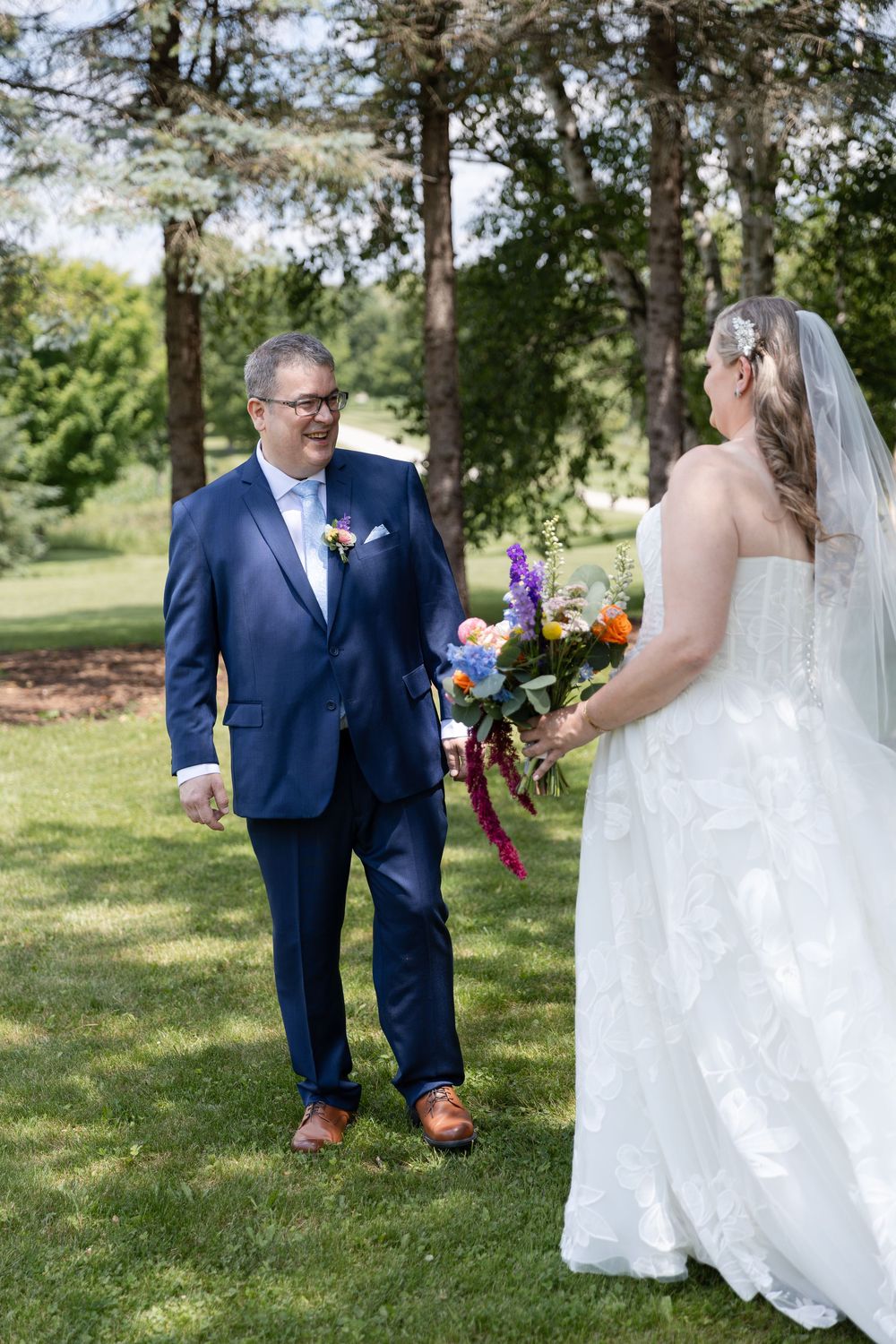 Groom in navy suit smiles at bride in white lace gown holding colorful bouquet during outdoor first look in Guelph, Ontario.