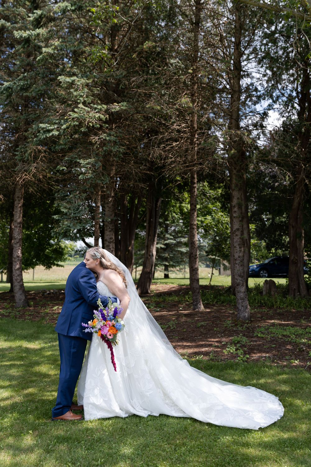 Bride and groom share a romantic kiss outdoors on green grass, surrounded by tall trees, bride holding colorful bouquet in Guelph, Ontario.