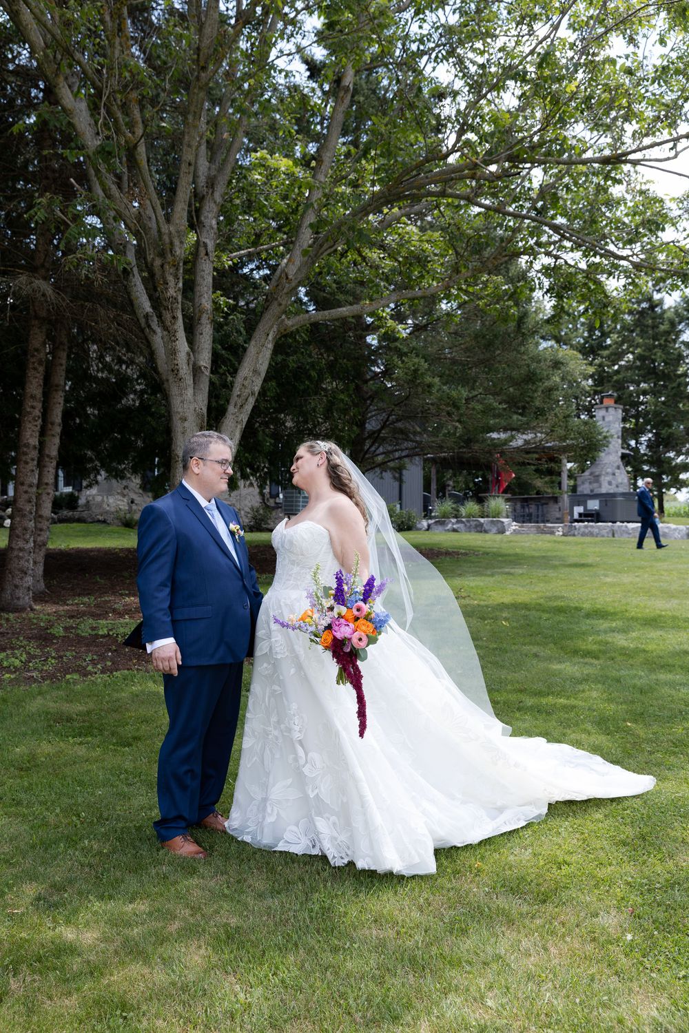 Bride in white lace gown holding colorful bouquet stands with groom in blue suit on green lawn with trees in Guelph, Ontario.