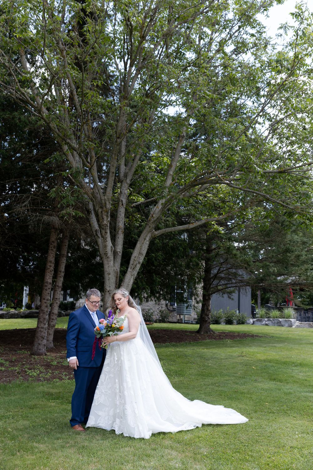 Bride in white lace gown with long train stands with suited man holding colorful bouquet on green lawn with trees in Guelph, Ontario.