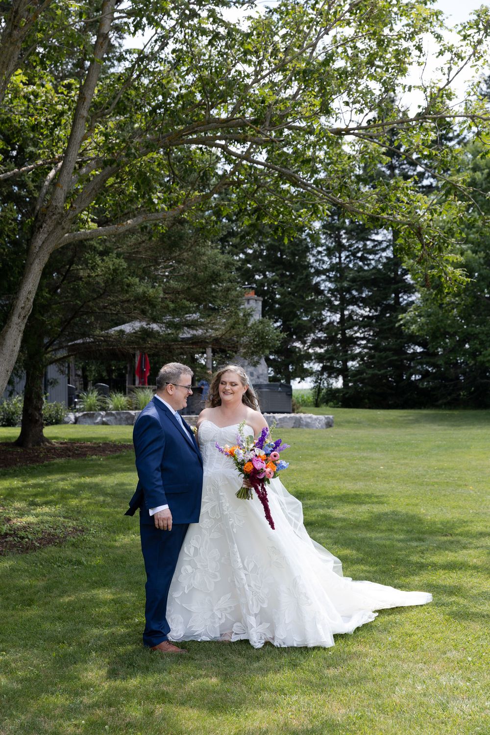 Bride in white lace gown holding colorful bouquet stands with groom in blue suit on green lawn with trees in Guelph, Ontario.