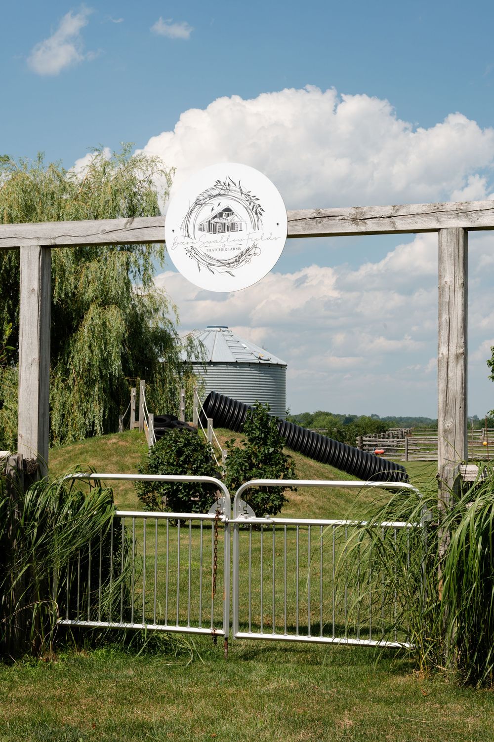 Rustic farm entrance with wooden pergola, metal gate, grain silo, and weeping willow trees under blue sky at barn swallow fields in guelph,