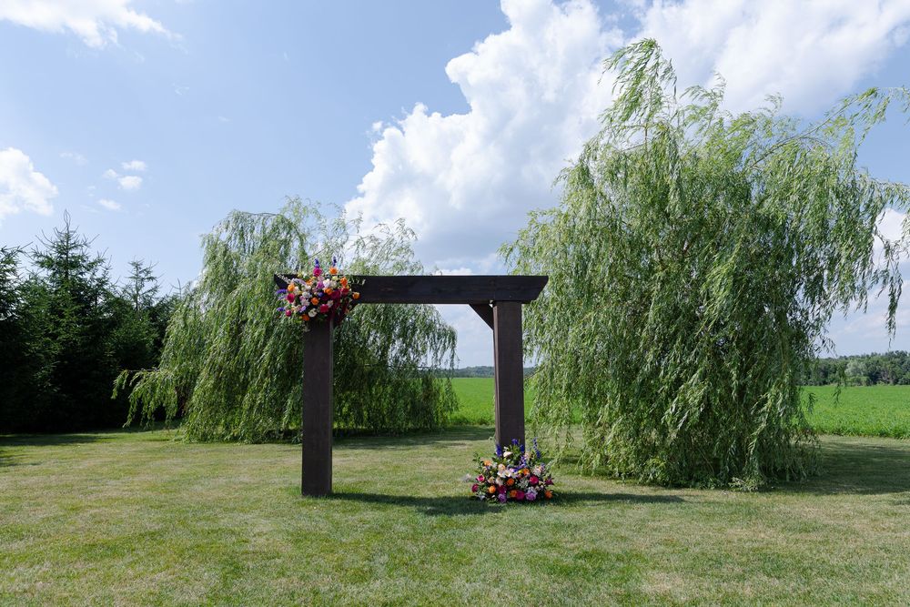 Rustic wooden wedding arch with colorful flowers on a lush green lawn with weeping willow trees at barn swallow fields in guelph, ontario