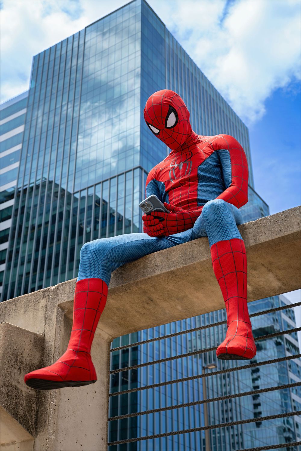 Person in Spider-Man costume sitting on concrete ledge checking phone with glass skyscraper in background.