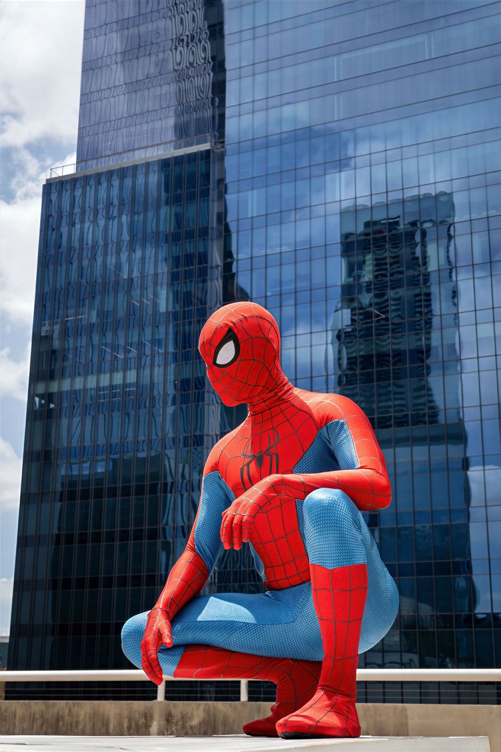Spider-Man costumed figure crouching heroically in front of a modern glass skyscraper on a sunny day.