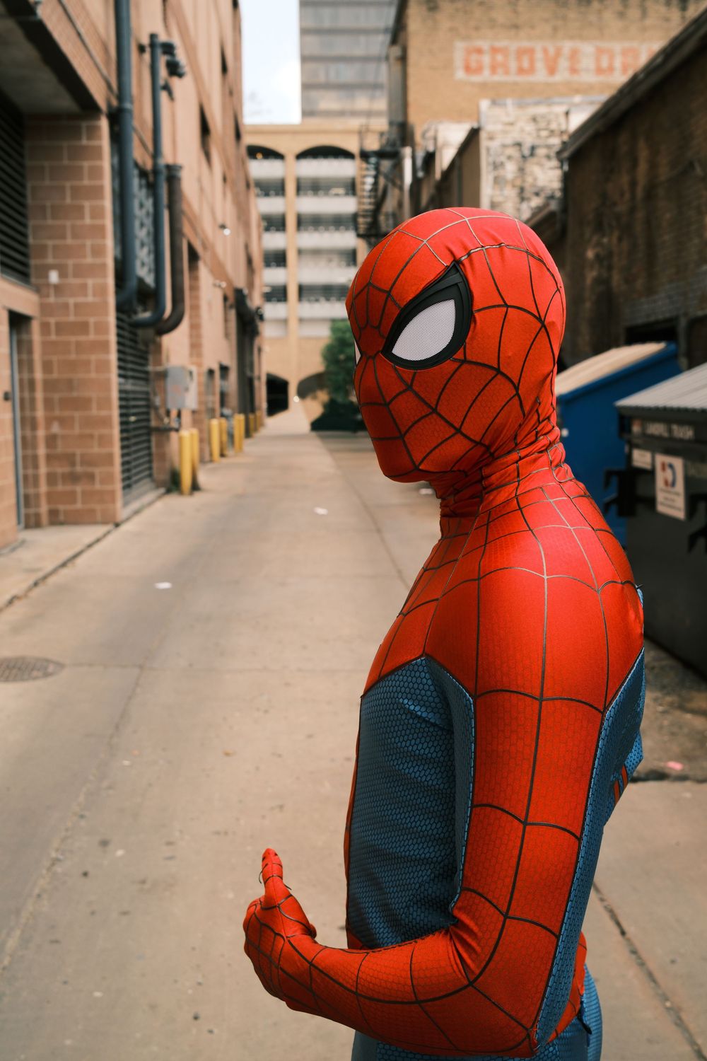 Person in a red and blue Spider-Man costume giving a thumbs up in an urban alley between brick buildings.