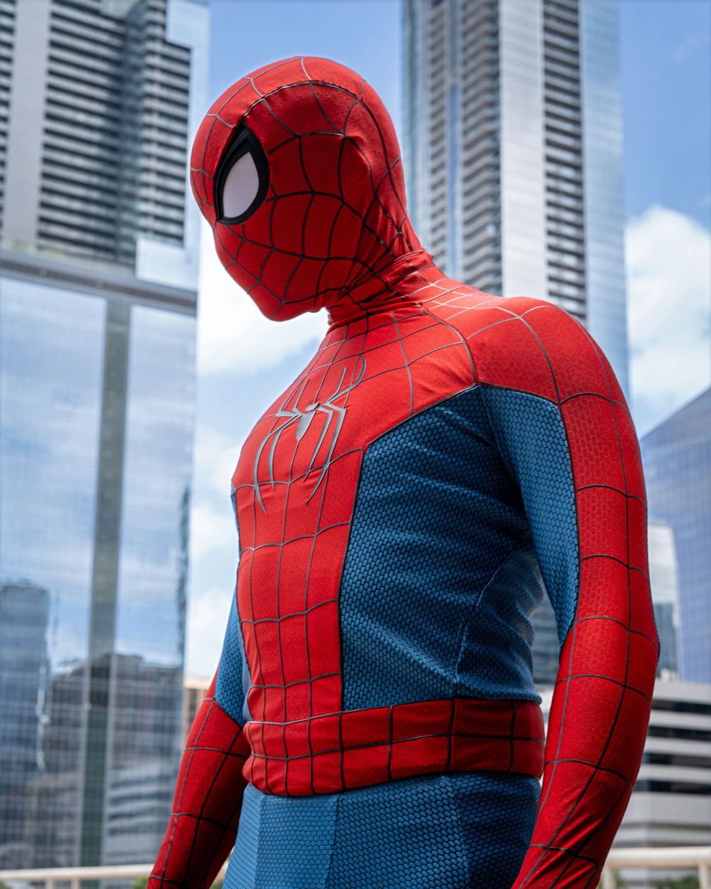 Person in a detailed red and blue Spider-Man costume posing in front of city skyscrapers on a sunny day.