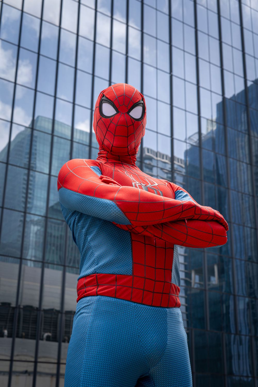 Person in a red and blue Spider-Man costume posing with arms crossed in front of a glass building.