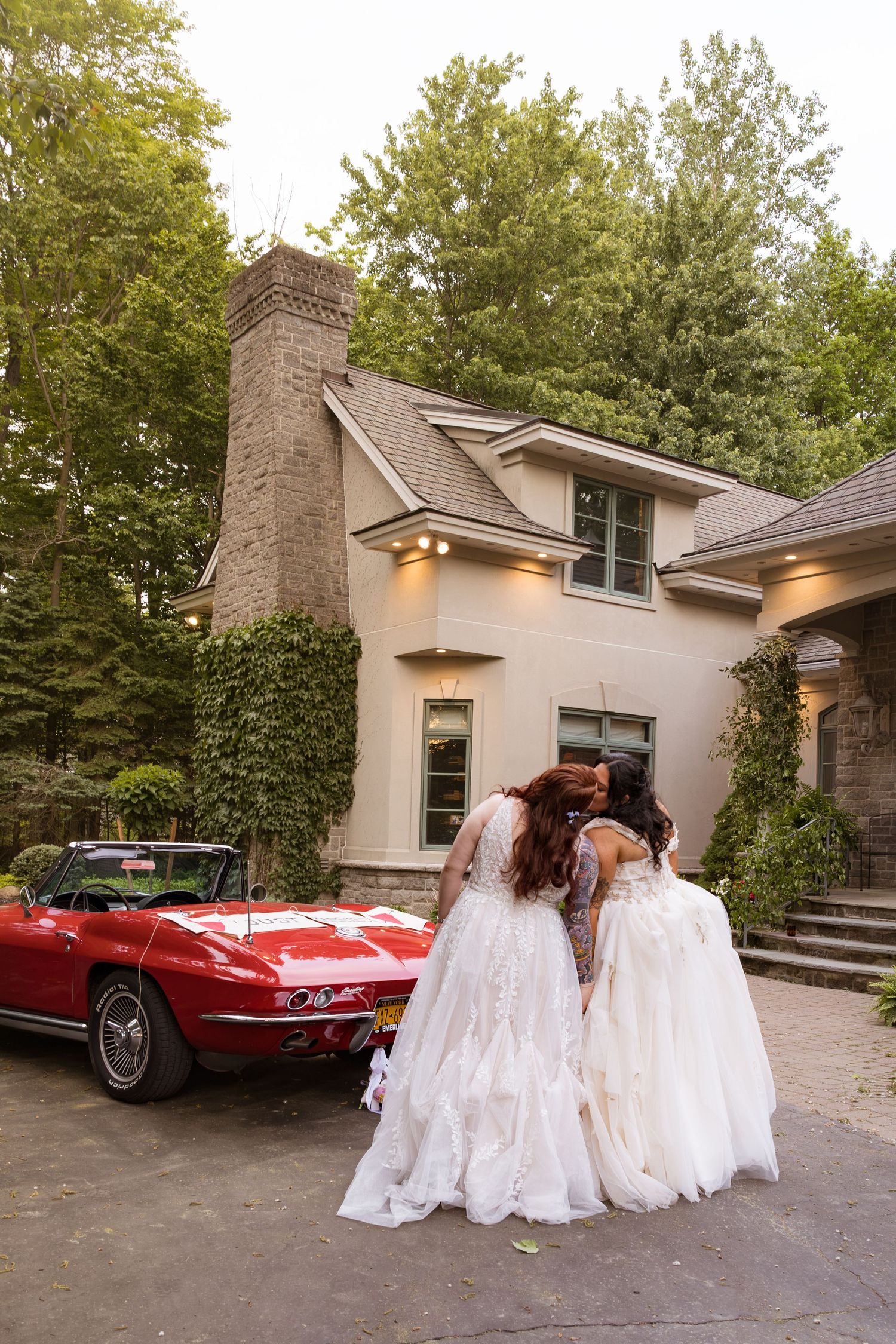 Two brides in white gowns embrace near a classic red Corvette outside an elegant home surrounded by trees.