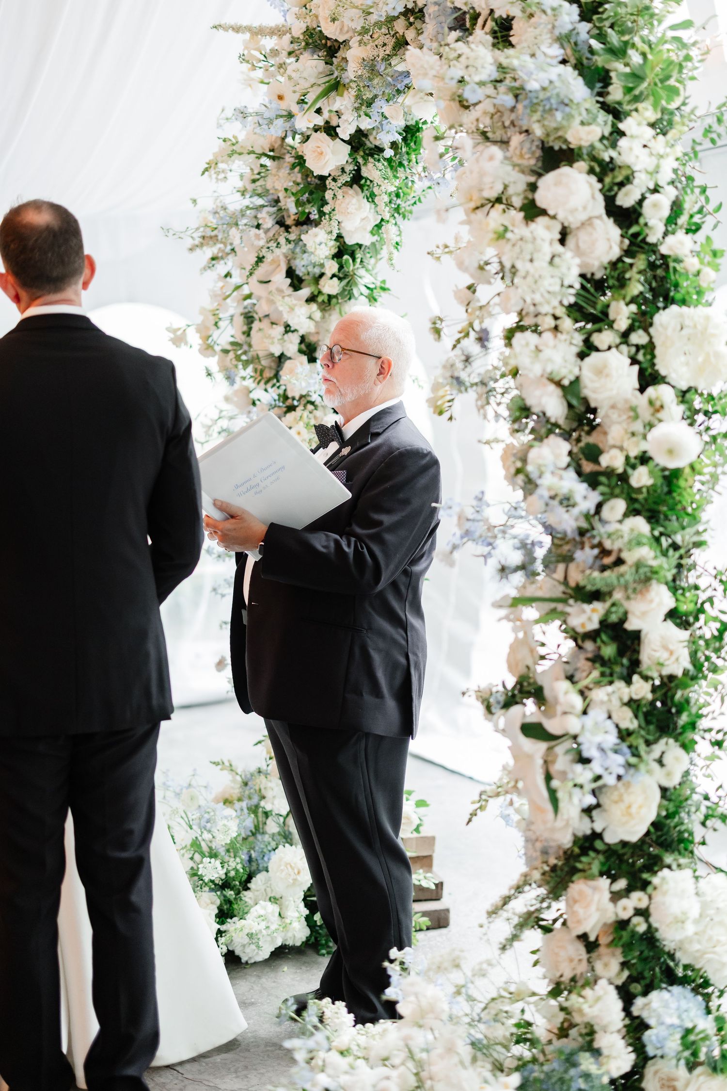 Officiant reads from ceremony booklet beside lush white and blue floral arch at wedding altar.